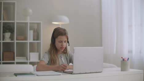 little-girl-is-learning-online-communicating-with-teacher-by-internet-using-headphones-and-laptop-doing-homework-in-exercise-book
